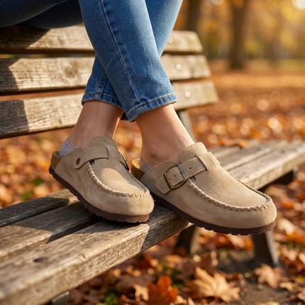 Womens suede clogs seated on a wooden bench with ankles crossed