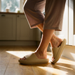 Women's cushioned slide sandals worn on a wooden kitchen floor in low morning sun