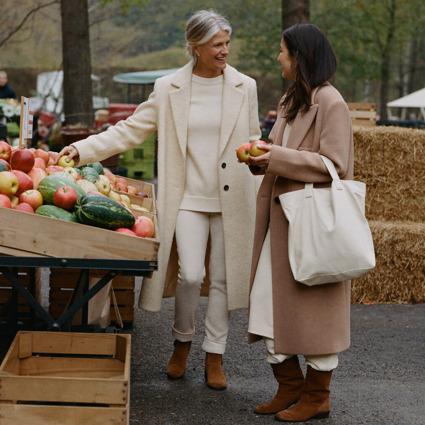 A mother and daughter shopping together at an autumn farmers market, both in cream knitwear and brown suede boots